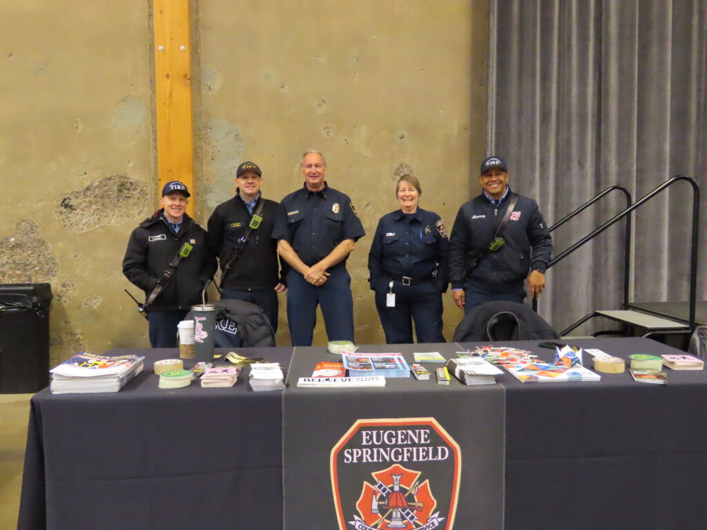 Five firefighters from the Eugene Springfield Fire Department stand behind a table filled with handouts.
