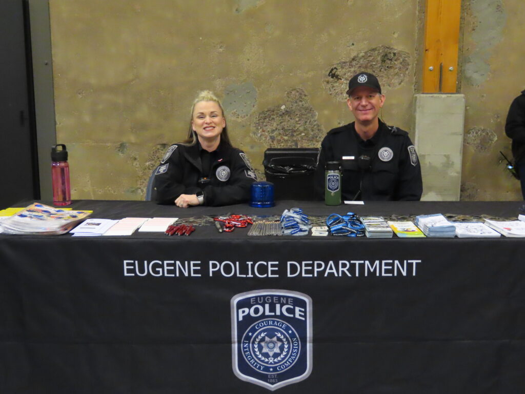 Two police officers from the Eugene Police Department sit behind a table filled with handouts. 