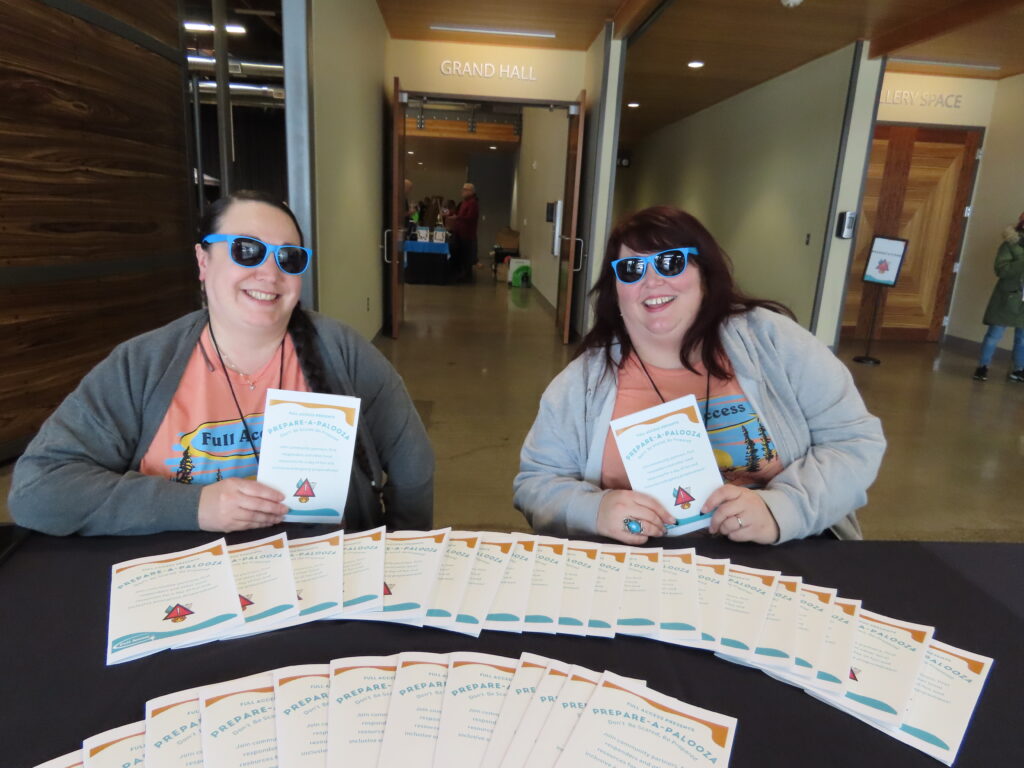 Two full access employees wearing fun sunglasses sit behind a table filled with Prepare-a-Palooza programs.